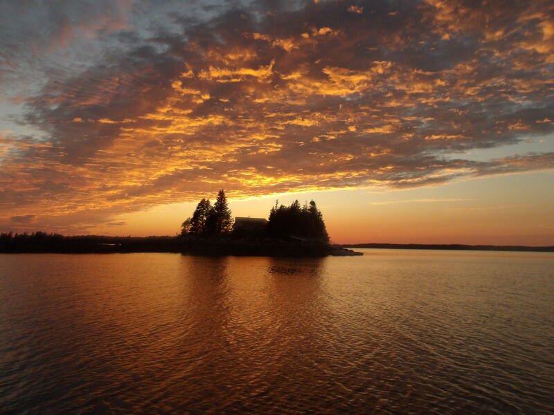 The image captures a serene sunset over a calm lake. The sky is ablaze with vibrant orange and yellow hues, reflecting on the water's surface. A small island with silhouetted trees sits in the distance, adding a touch of mystery to the scene. The overall mood is peaceful and picturesque.